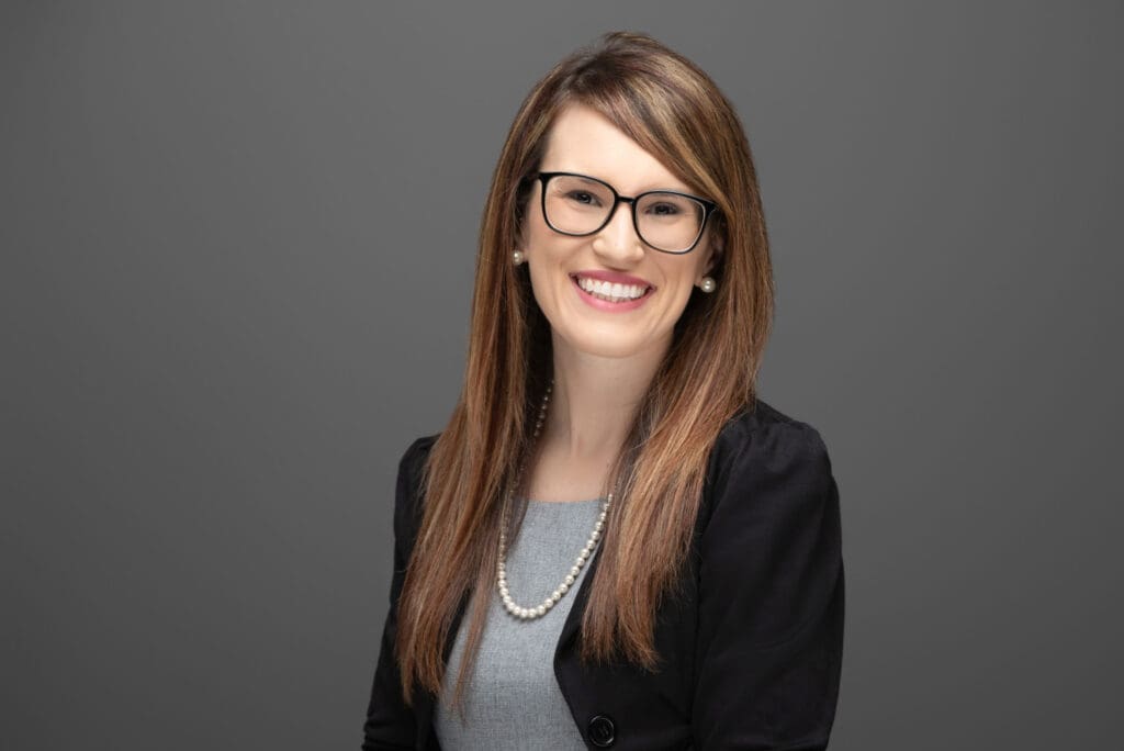 Professional executive headshot of a smiling woman wearing glasses and a black blazer against a gray studio background in Cincinnati, Ohio.