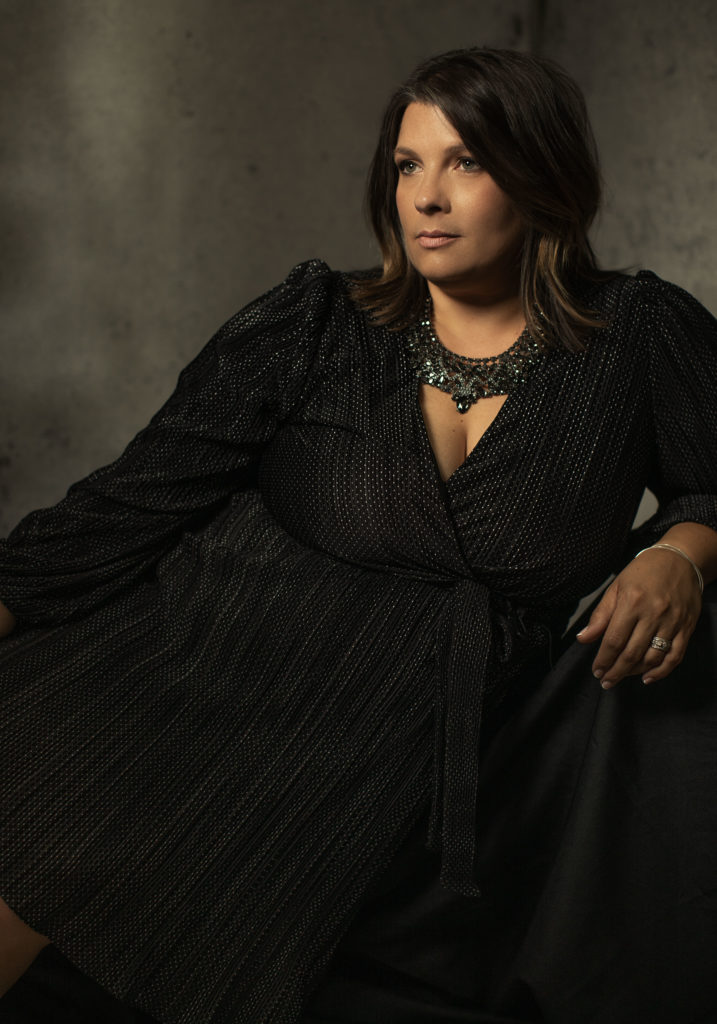 A woman reclines a looks off in the distance. She wears a black dress and a necklace. The purpose of the photo is for the 40 over 40 portrait project. 