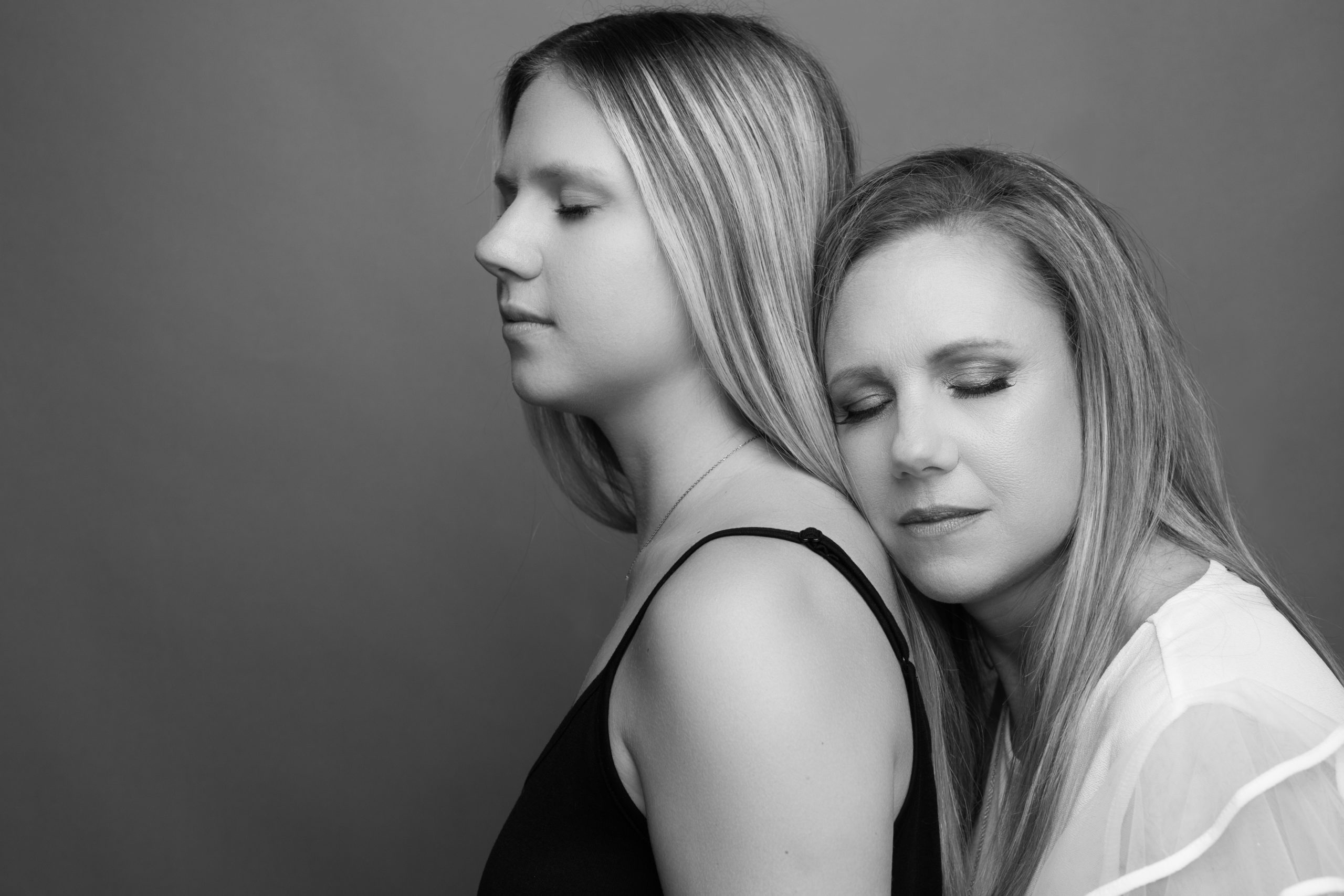 A mother rests her head on her adult daughter's back as they pose for a black and white image. Both women have their eyes closed.