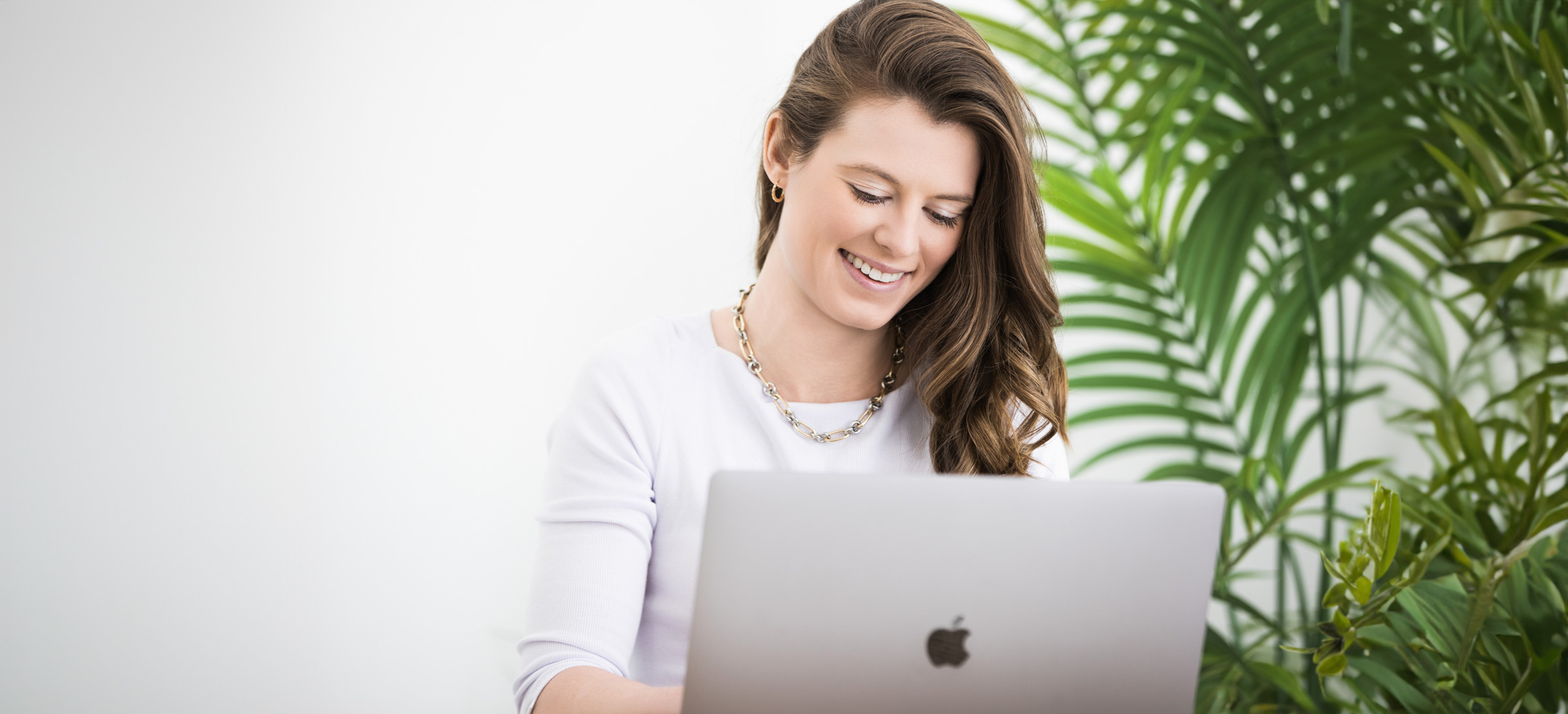 Professional headshot of a smiling woman in a white top, working on a laptop with a bright, clean backdrop and green plant. Captured by Janel Lee Photography in Cincinnati, Ohio – specializing in modern headshots and personal branding portraits for professionals.