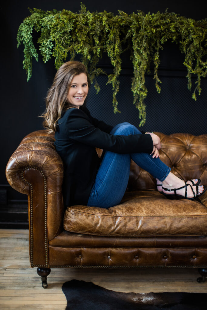 Relaxed yet professional portrait of a woman in a black blazer and jeans, seated on a leather couch with a moody green plant backdrop. Janel Lee Photography in Cincinnati specializes in contemporary personal branding photography for professionals.