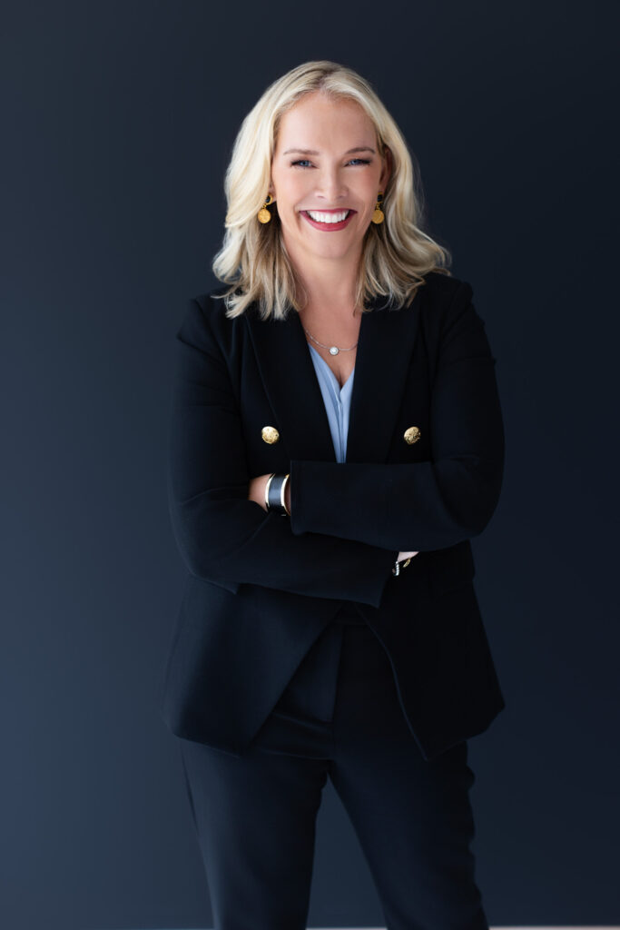 Cathy Major exudes confidence in a professional branding portrait taken at Janel Lee Photography in Cincinnati, Ohio. She stands with arms crossed, wearing a sophisticated black blazer with gold button accents, paired with a soft blue blouse. The deep, moody backdrop enhances her polished and authoritative presence, making this the perfect image for a high-end luxury travel professional.