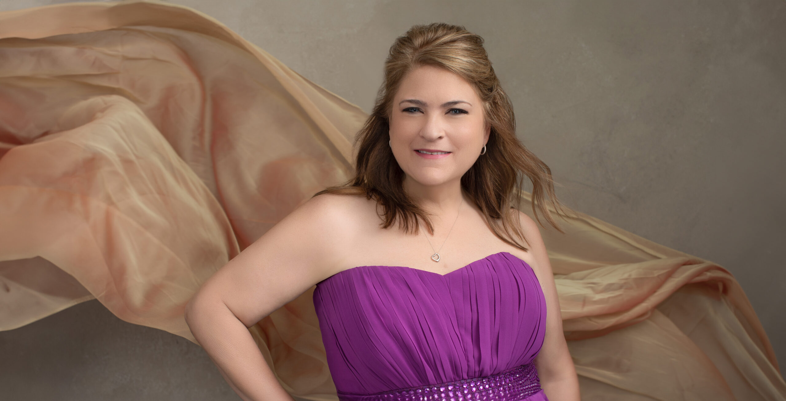 A vibrant personal branding portrait of a Cincinnati woman in a strapless purple gown with sparkling details at the waist. A soft, flowing peach-colored fabric cascades in the background, adding elegance and movement. This empowering image is ideal for women in the Cincinnati area looking for glamorous and sophisticated portraits.