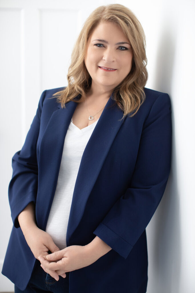 A personal branding portrait of a Cincinnati professional woman leaning casually against a white wall in a downtown photography studio. She wears a navy blazer over a white top, with her hands gently clasped. Her warm smile and confident demeanor make this headshot perfect for LinkedIn or other professional profiles in the Cincinnati area.