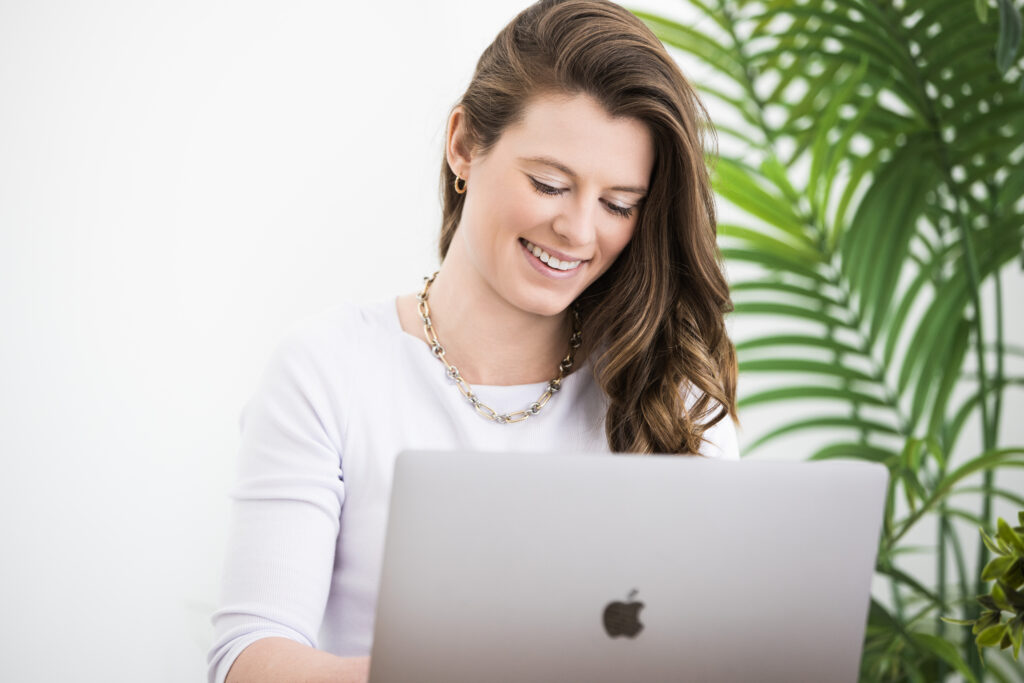 Professional headshot of a smiling woman in a white top, working on a laptop with a bright, clean backdrop and green plant. Captured by Janel Lee Photography in Cincinnati, Ohio – specializing in modern headshots and personal branding portraits for professionals.