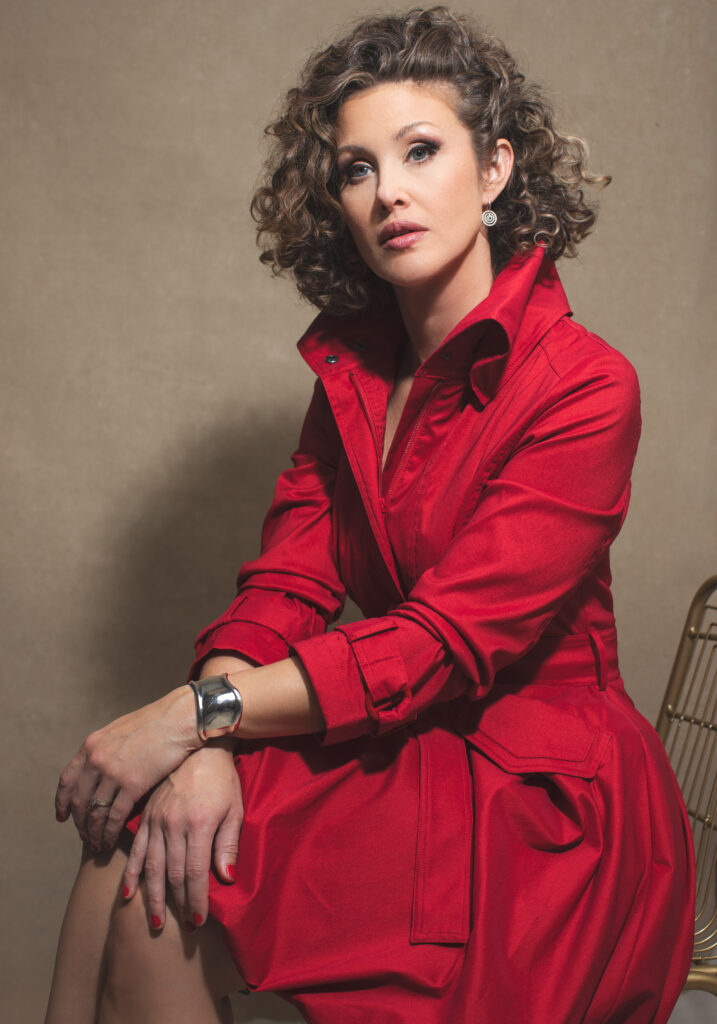 A Cincinnati-based portrait of a woman with curly hair seated confidently in a red trench dress against a neutral beige background. Photographed in a professional studio, she exudes elegance with her silver cuff bracelet, red nails, and delicate earrings. This stunning portrait captures timeless beauty, ideal for personal branding or headshots.