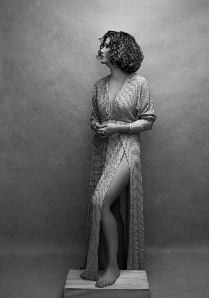 A Cincinnati studio portrait of a woman standing barefoot on a wooden block against a textured gray background. She wears a flowing wrap dress with a high slit, her profile turned slightly for a graceful and timeless composition. This portrait is perfect for women in Cincinnati seeking classic, empowering images.