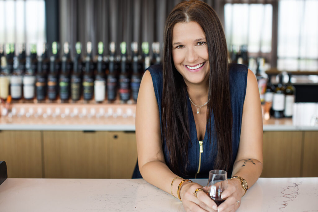 Cincinnati professional branding portrait of a confident woman with long brown hair leaning on a marble bar, smiling warmly while holding a bourbon tasting glass. Photographed at New Riff Distilling by Janel Lee Photography, with a blurred background of premium bourbon bottles creating a polished and approachable atmosphere for corporate and personal branding sessions.
