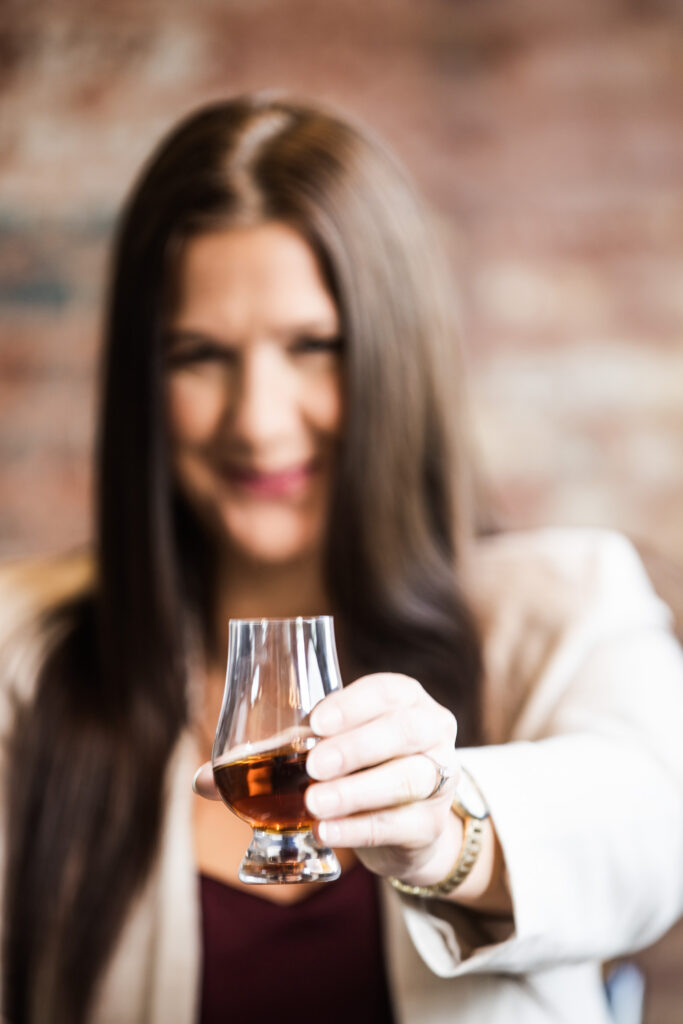 Creative branding portrait of a woman with long straight brown hair holding a Glencairn glass of bourbon toward the camera, smiling softly with a blurred brick wall backdrop. Captured at Janel Lee Photography’s Cincinnati studio, this image reflects a confident and engaging personal brand for professionals in the bourbon or sales industry.