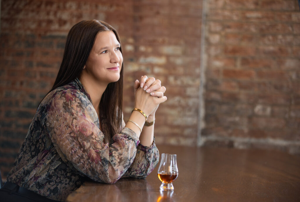 Professional personal branding portrait of a confident woman with long brown hair seated at a wooden table, smiling gently with hands clasped, wearing a sheer floral blouse and gold bracelets. Captured at Janel Lee Photography’s downtown Cincinnati studio with a warm, exposed brick backdrop and a glass of bourbon on the table, reflecting a sophisticated and approachable brand image.