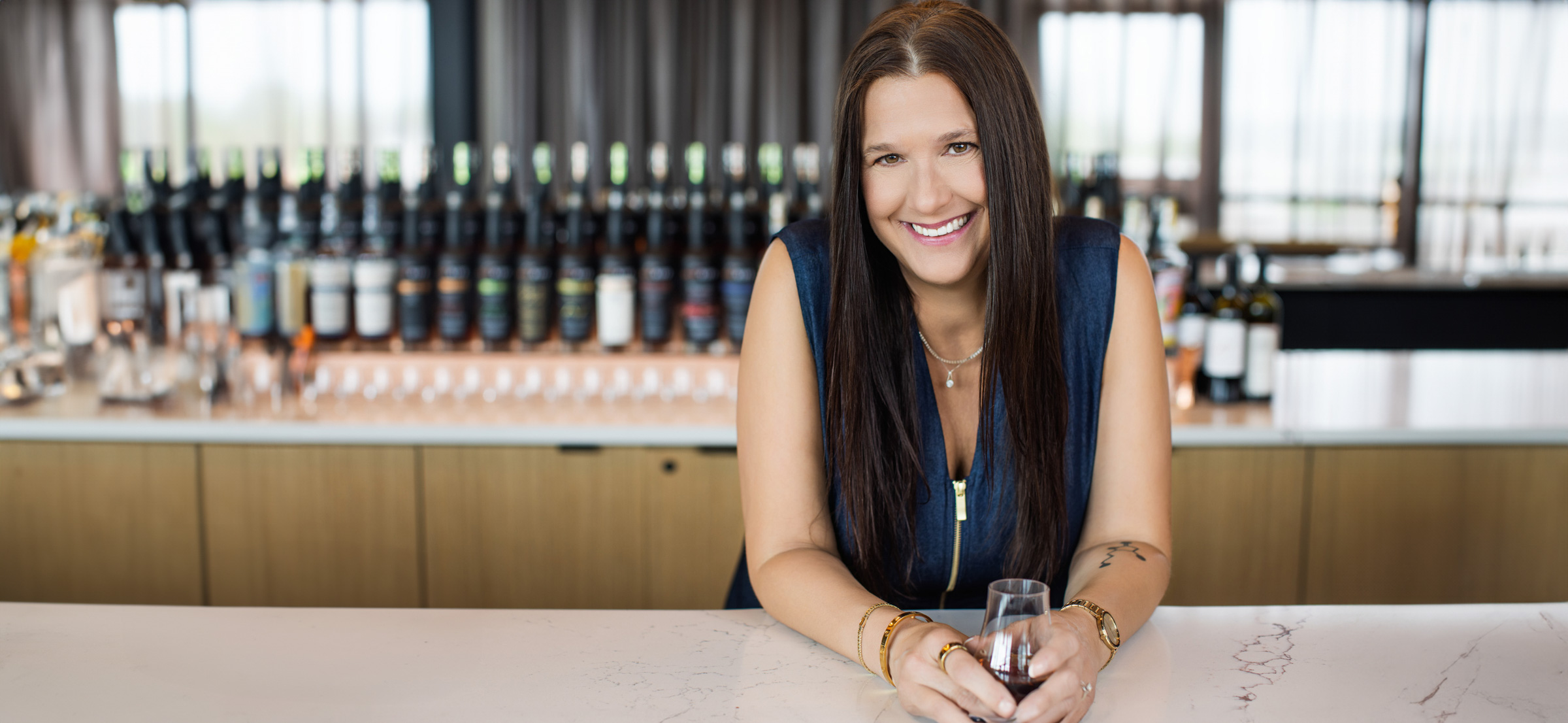 Cincinnati professional branding portrait of a confident woman with long brown hair leaning on a marble bar, smiling warmly while holding a bourbon tasting glass. Photographed at New Riff Distilling by Janel Lee Photography, with a blurred background of premium bourbon bottles creating a polished and approachable atmosphere for corporate and personal branding sessions.