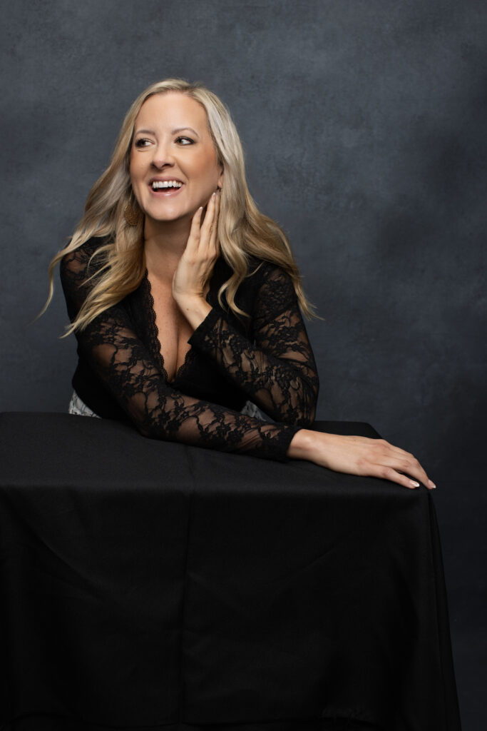 Katie Stuart smiling at black table in lace top with dark studio background