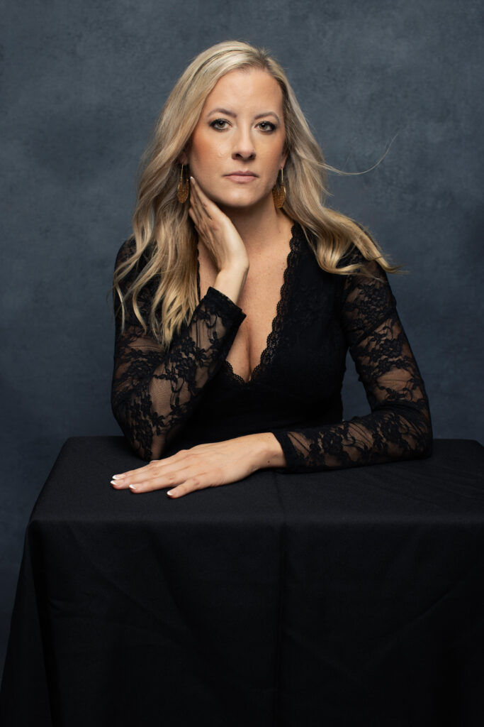Katie Stuart seated at black table wearing lace top in dark studio portrait