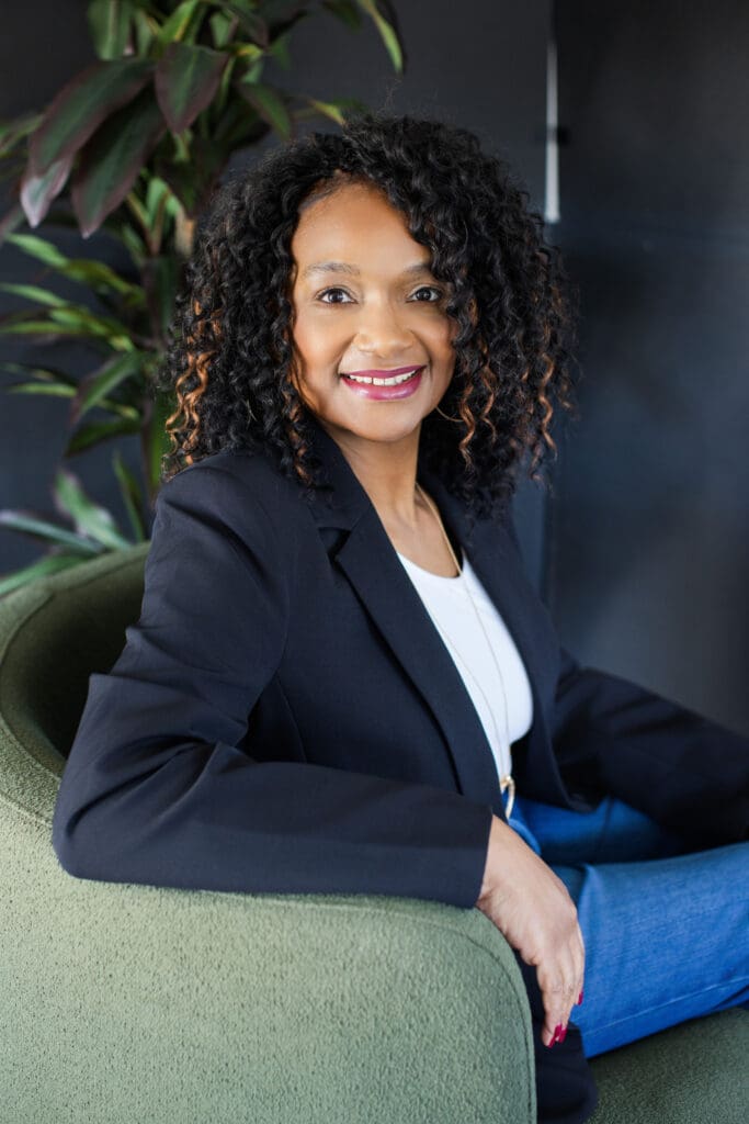 Sheila Eason seated in green chair, smiling in blazer and jeans
