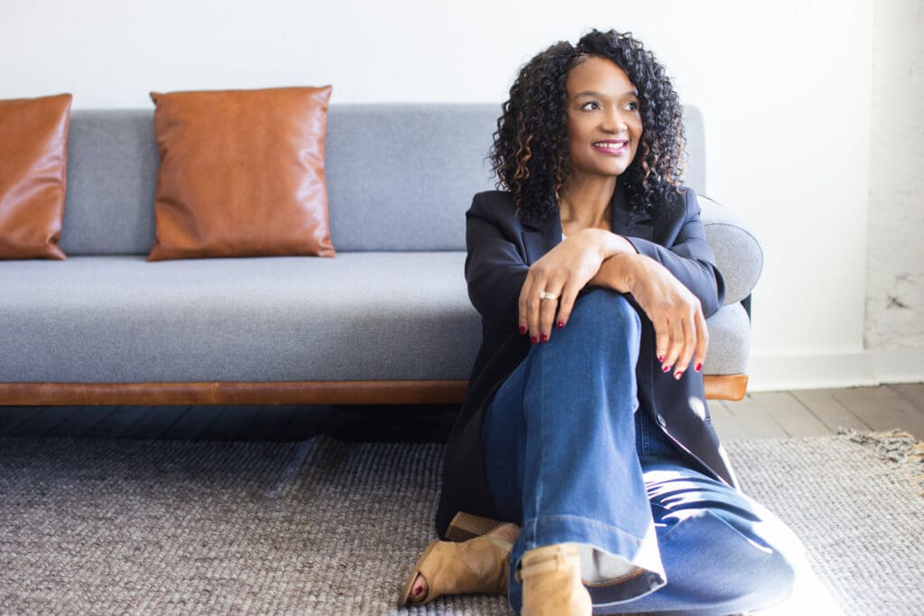Sheila Eason sitting on the floor in casual jeans and blazer, smiling