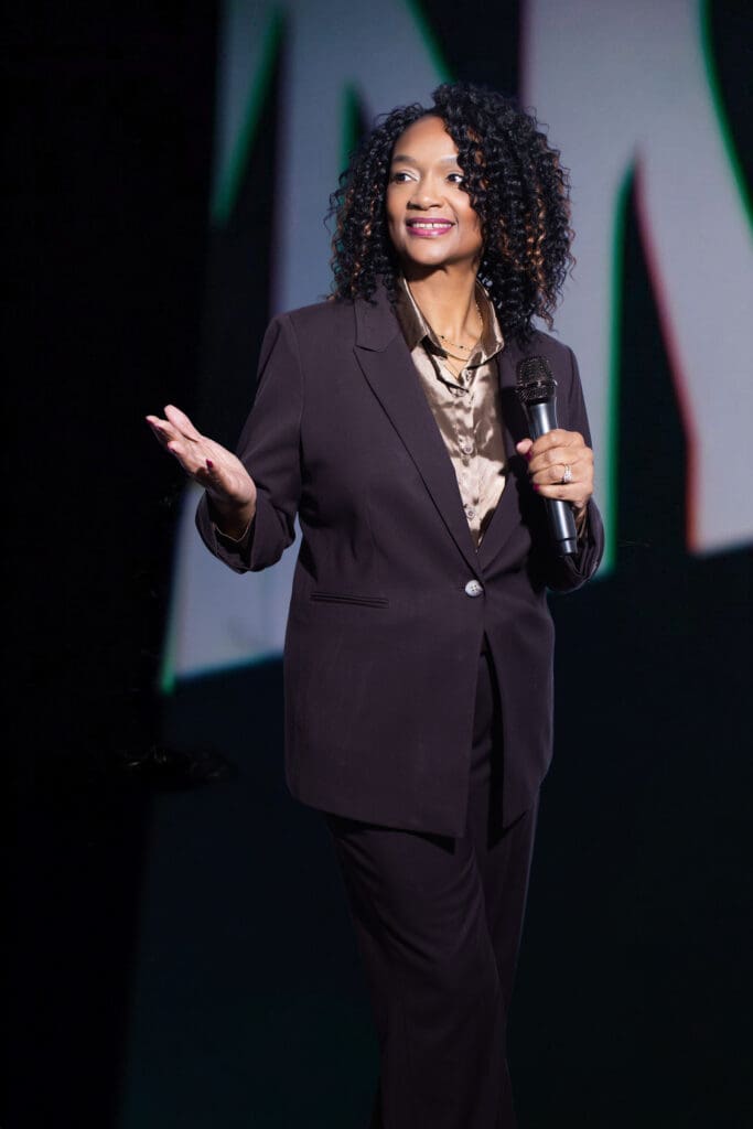 Sheila Eason speaking on stage in a dark suit with microphone in hand
