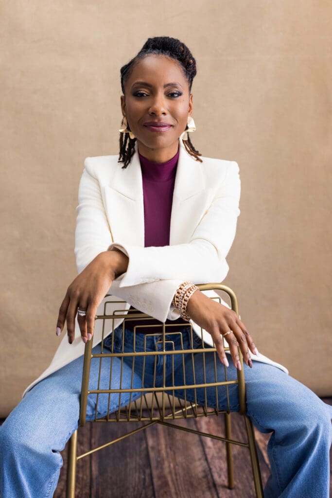 Professional personal branding portrait of a confident woman photographed in a Cincinnati portrait studio, seated backwards on a modern metal chair with her arms resting casually across the chair back. She wears a tailored cream blazer layered over a plum high-neck top and relaxed denim, blending polished professionalism with approachability. Her expression is calm, self-assured, and direct, conveying confidence, leadership, and credibility. Gold statement earrings, stacked bracelets, and a ring add subtle elegance. Her hair is styled in neat locs pulled back, and the neutral, softly textured studio backdrop paired with warm wood flooring creates a refined, editorial environment ideal for executive headshots, LinkedIn profile photos, and personal branding photography in Cincinnati, Ohio.
