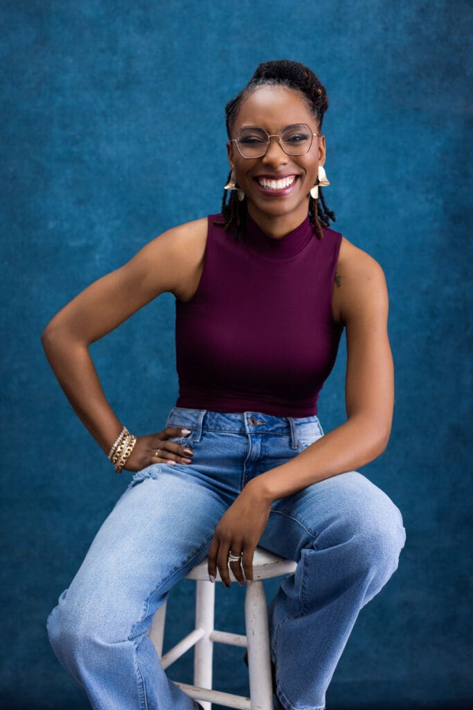 Modern personal branding portrait of a confident, smiling woman photographed in a professional Cincinnati portrait studio against a rich blue textured backdrop. She is seated casually on a studio stool with one hand resting on her hip and the other relaxed on her knee, creating an open, self-assured pose. She wears a sleeveless plum top paired with relaxed denim and clear-framed glasses, blending approachability with professionalism. Gold statement earrings, stacked bracelets, and a ring add subtle polish. Her hair is styled in neat locs pulled back, and her bright, natural smile conveys confidence, warmth, and authenticity. The clean studio lighting and bold yet refined backdrop make this image ideal for personal branding photography, LinkedIn profiles, and professional marketing imagery in Cincinnati, Ohio.