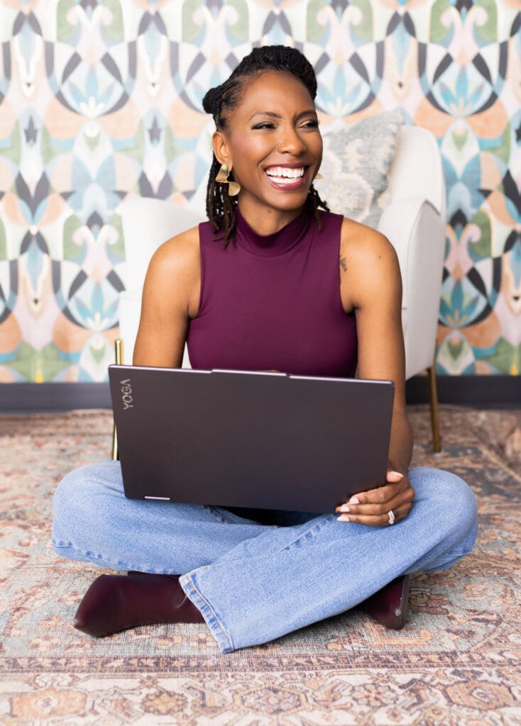 Lifestyle personal branding portrait of a confident, smiling woman photographed in a professional Cincinnati portrait studio. She is seated cross-legged on a patterned rug, working comfortably on a laptop, creating a relaxed and modern professional feel. She wears a sleeveless plum top paired with jeans and coordinating accessories, including gold statement earrings and a ring, blending approachability with polish. Her hair is styled in neat locs pulled back, and her bright, genuine smile conveys warmth, confidence, and ease. The backdrop features a softly lit studio setting with a white accent chair and a colorful, geometric wallpaper, adding visual interest while maintaining a clean, contemporary aesthetic. This image is ideal for personal branding photography, LinkedIn profiles, and professional marketing imagery in Cincinnati, Ohio.