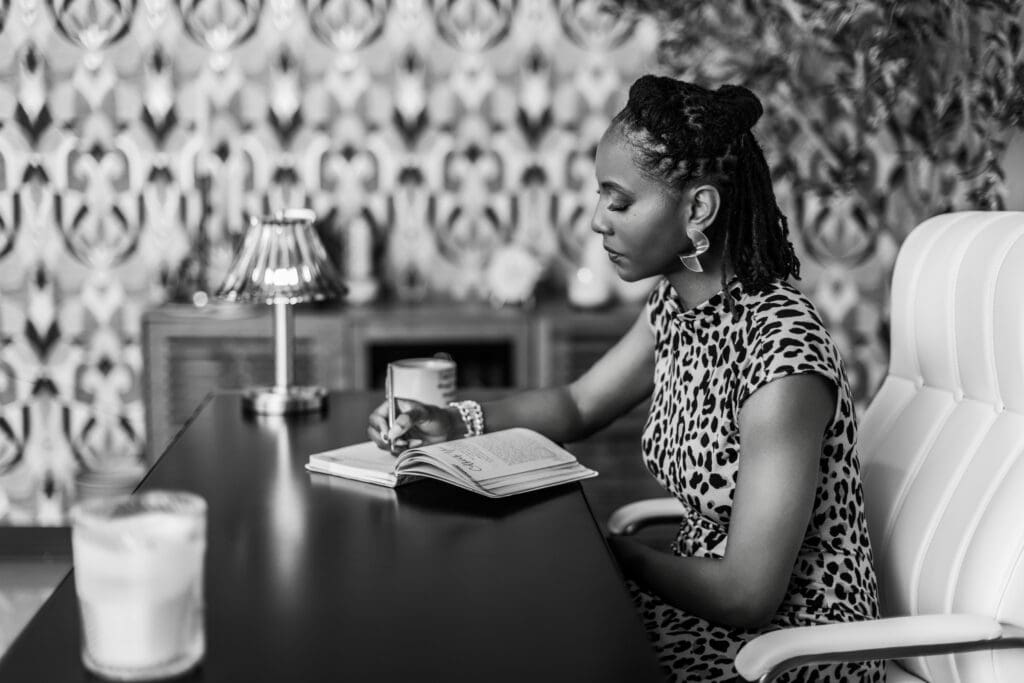 Black-and-white lifestyle personal branding portrait of a focused, confident woman photographed in a professional Cincinnati portrait studio. She is seated at a desk in profile, thoughtfully writing in an open notebook while holding a pen, conveying concentration, intention, and leadership. She wears a sleeveless patterned dress with gold statement earrings and stacked bracelets, blending professionalism with personal style. Her hair is styled in neat locs pulled back, and her calm, composed expression reflects clarity and purpose. The setting features a refined studio environment with a modern desk, a soft table lamp, a white office chair, and a decorative patterned wall in the background, creating an editorial, story-driven atmosphere. This image is well suited for personal branding photography, executive storytelling, and professional marketing imagery in Cincinnati, Ohio.