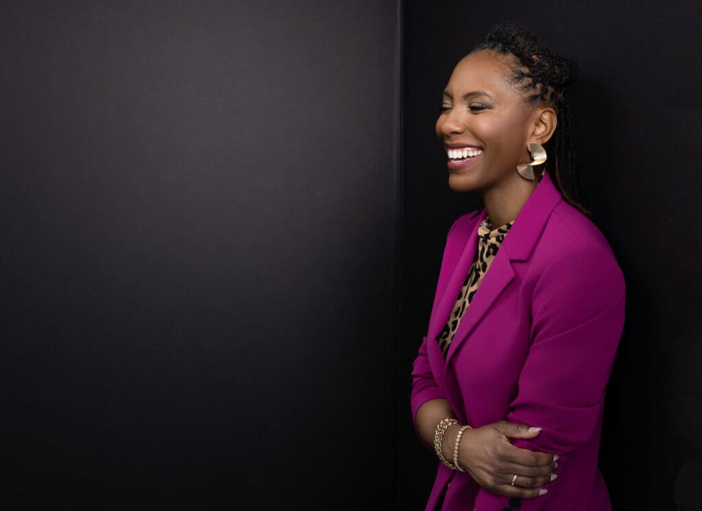 Side-profile personal branding portrait of a confident, joyful woman photographed in a professional Cincinnati portrait studio. She is turned slightly to the side, laughing naturally with her eyes gently closed, conveying warmth, authenticity, and ease. She wears a rich magenta blazer layered over a patterned blouse, paired with gold statement earrings and stacked bracelets. Her hair is styled in neat locs pulled back, and her relaxed posture with arms crossed adds to the candid, approachable feel. The backdrop is a deep, dark neutral background with subtle gradient tones, creating a dramatic, editorial look that keeps full focus on expression and presence. This image is well-suited for executive headshots, LinkedIn profile photos, and personal branding photography in Cincinnati, Ohio.