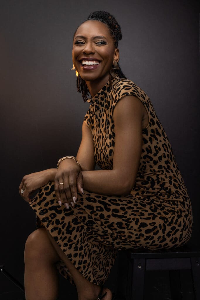Editorial-style personal branding portrait of a confident, smiling woman photographed in a professional Cincinnati portrait studio. She is seated on a stool in a relaxed three-quarter pose, leaning forward slightly with one arm resting on her knee, creating an open and approachable posture. She wears a sleeveless leopard-print dress paired with gold statement earrings, a bracelet, and a ring, adding personality and polish to the look. Her hair is styled in neat locs pulled back, and her wide, genuine smile conveys warmth, confidence, and ease. The backdrop is a dark, seamless studio background with soft, directional lighting that highlights her expression and skin tone while maintaining a refined, editorial feel. This image is well suited for executive portraits, LinkedIn profile photos, and personal branding photography in Cincinnati, Ohio.