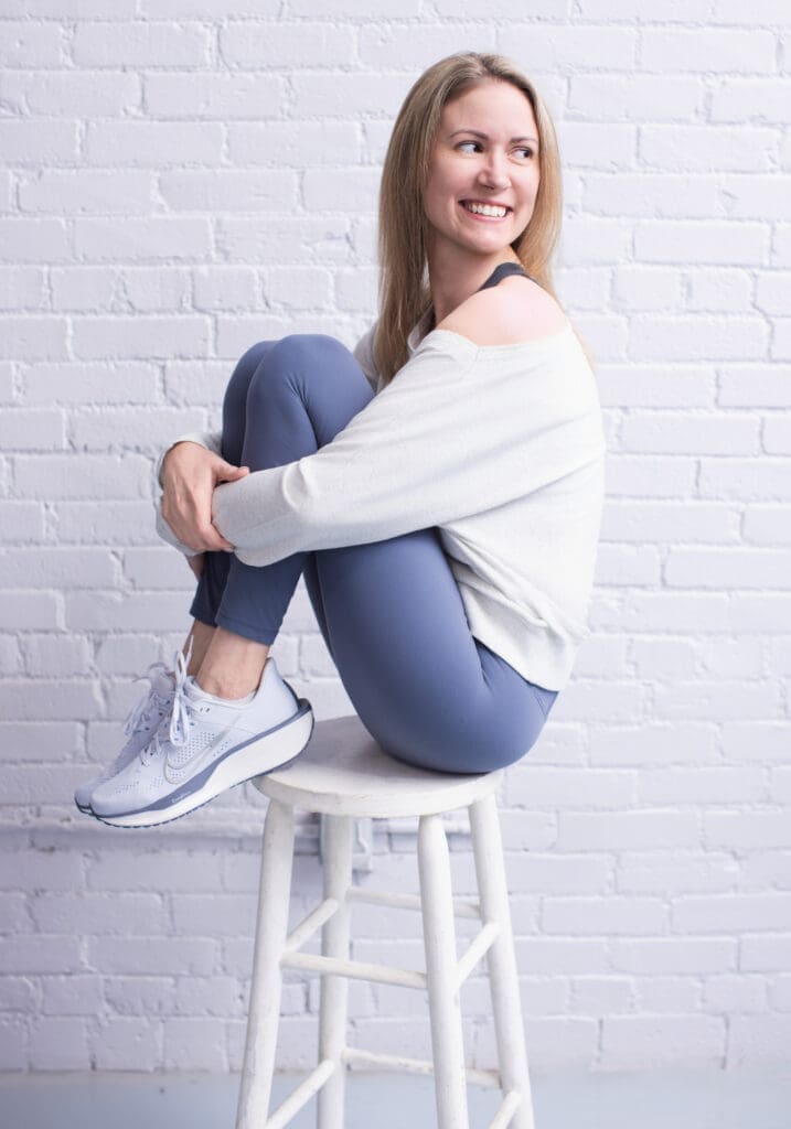 Portrait of 40 Over 40 participant Lynsey Gaca in Cincinnati, Ohio, smiling while seated on a white stool in casual athletic wear against a white brick studio wall.