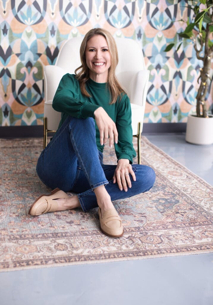 Portrait of 40 Over 40 participant Lynsey Gaca in Cincinnati, Ohio, smiling while seated on a patterned rug in a stylish studio with colorful wallpaper, wearing a green blouse and jeans.