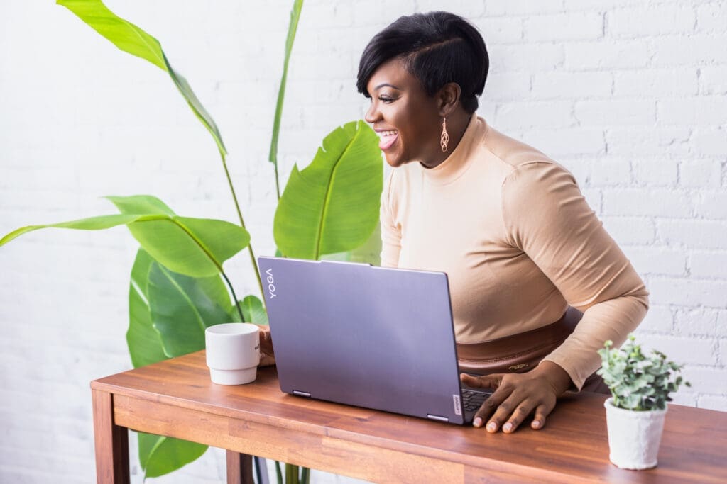 Personal branding photo of writer and speaker Paulette Jones in Cincinnati, Ohio, smiling while working on a laptop at a modern workspace with greenery and a bright studio setting.