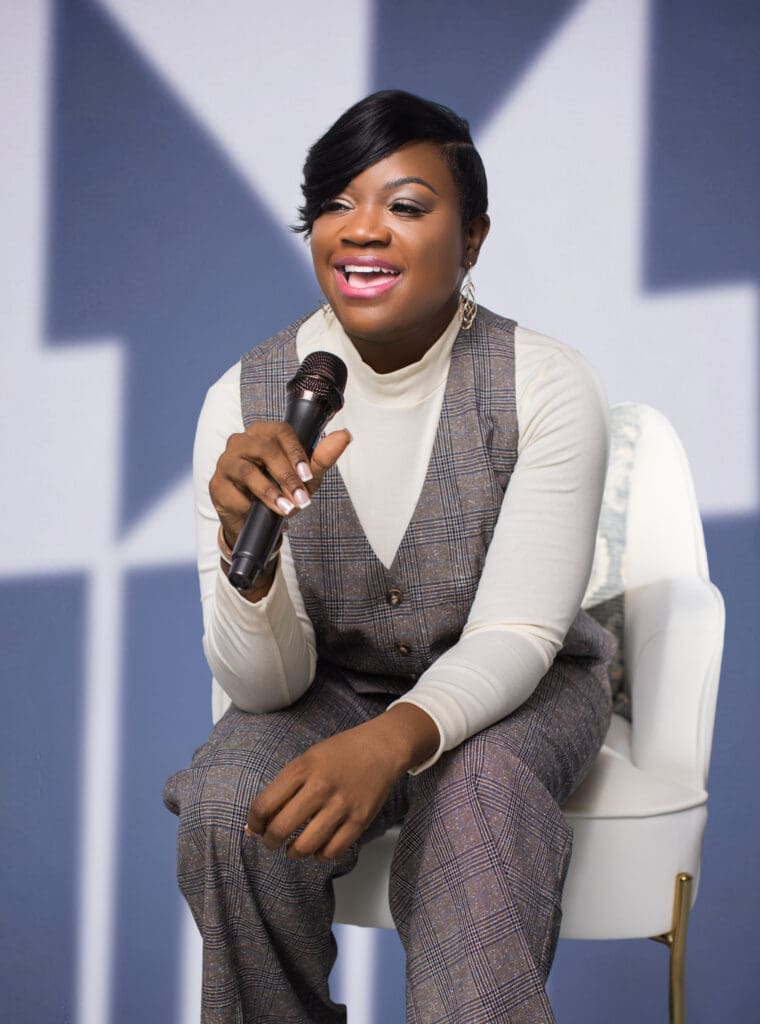 Personal branding portrait of writer and speaker Paulette Jones in Cincinnati, Ohio, smiling while holding a microphone and seated in a modern studio setting wearing a plaid jumpsuit and white turtleneck.